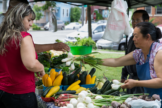 Two women exchange market tokens for green onions at a farmers market.