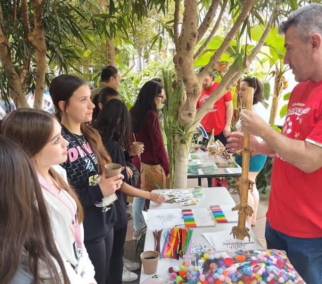 Two girls looking at demonstration from a male educator that is holding a stick and craft items on a table in front of him