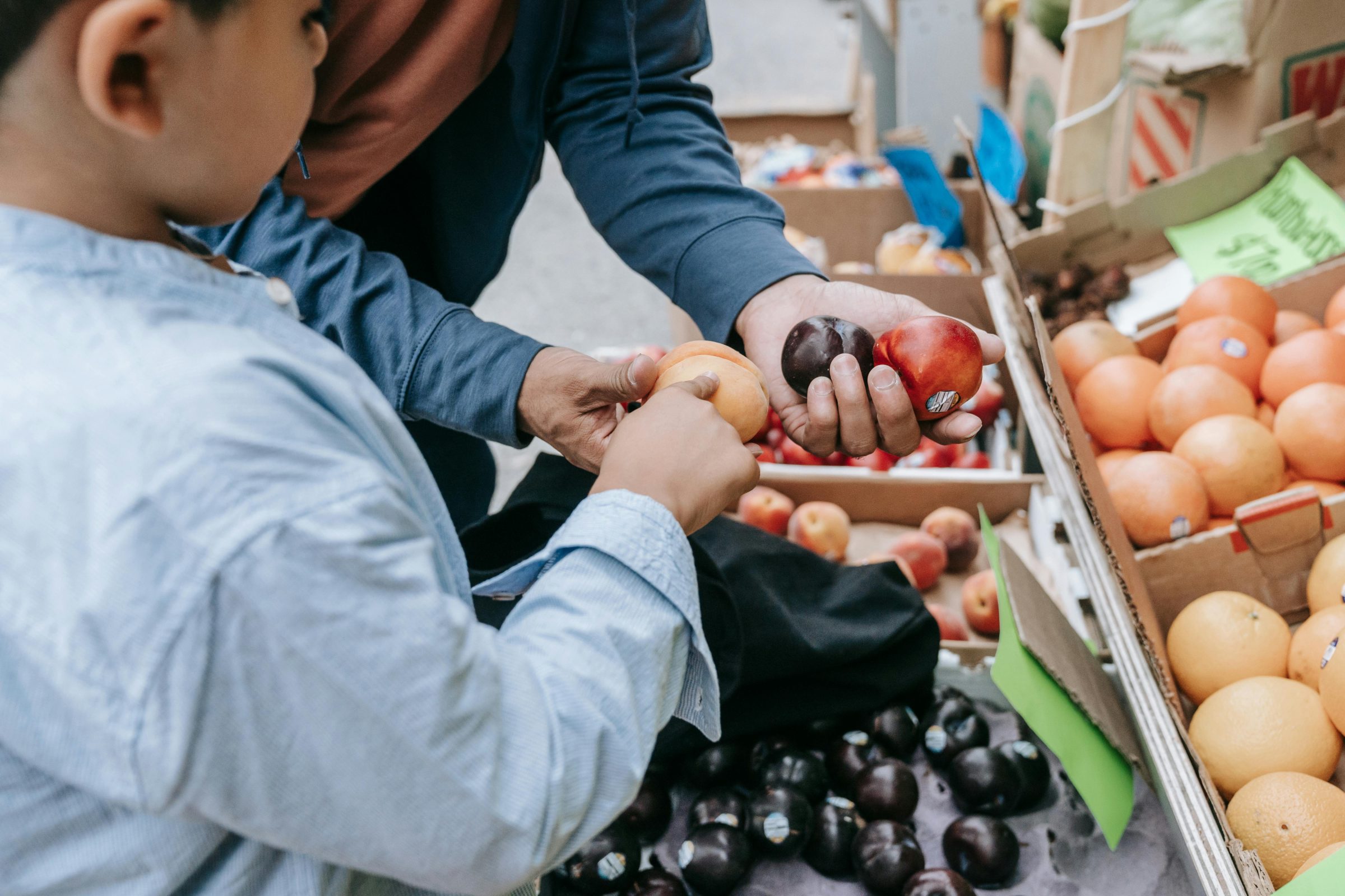 hands of an adult and a half of a child's face both reaching to touch peaches and plums in a market